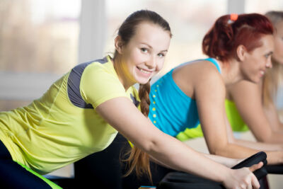 Fitness practice, group of beautiful cheerful females cycling in sports club, doing cardio exercises for slimming, warming up during sport lesson in class, focus on smiling woman with braid