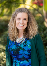 Woman in a blue and green floral top with a green sweater in front of greenery.