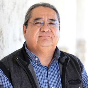 Native American man in blue dress shirt with a black vest in front of a white background.