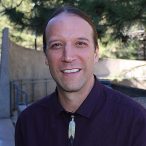 Native American man in a black shirt in front of a greenery and concrete background.